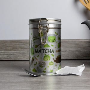 Puede incluir: Un bote de té matcha de metal plateado con tapa abisagrada y cierre. El bote está decorado con ilustraciones verdes de artículos relacionados con el matcha y la palabra "Matcha". Una cuchara y un pequeño paquete de papel están sobre la superficie de madera.