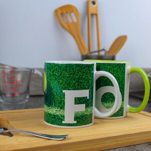 Könnte beinhalten: Zwei weiße Keramikbecher mit grünen und weißen Designs. Die Becher haben ein grünes und weißes Grasdesign mit einem weißen Buchstaben "F" auf einem Becher. Die Becher stehen auf einem Holzbrett.