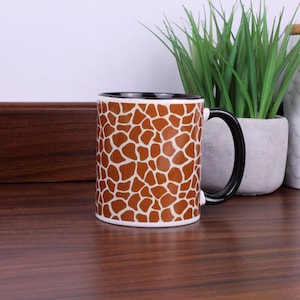 May include: A ceramic mug featuring a brown and white giraffe print pattern across the exterior surface. The mug has a solid black handle and a black interior rim, providing a distinct contrast to the animal-inspired design on the body.
