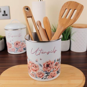 May include: White utensil pot with a floral design and the word "Utensils" in pink script. It holds wooden kitchen tools, including a spatula and tongs. A matching biscuit jar is visible in the background.