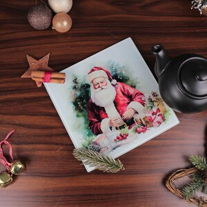 Può includere: Piatto decorativo quadrato a tema natalizio con Babbo Natale in abito rosso, barba bianca e cappello. Il piatto è circondato da decorazioni natalizie, una stella, campane e una teiera nera su una superficie di legno.