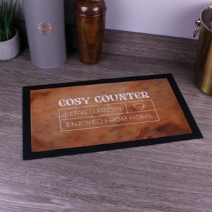 Può includere: Un tappetino rettangolare con bordo nero e sfondo marrone maculato. Il testo recita COSY COUNTER, SERVED FRESH e ENJOYED FROM HOME, con una piccola icona a linee di una tazzina da caffè. Il design è centrato sul tappetino dalla superficie piatta.