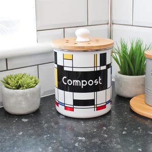 Könnte beinhalten: Ein weißer Komposteimer mit Bambusdeckel und weißem Knauf. Der Eimer hat ein schwarzes Rechteck mit dem Wort "Compost" in Weiß und ein farbenfrohes geometrisches Design mit gelben, blauen und roten Akzenten.