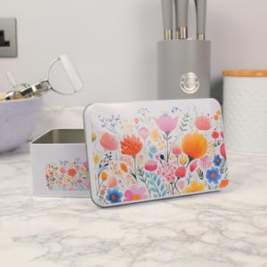 Op de afbeelding: Een witte rechthoekige blikken doos met een kleurrijk bloemenontwerp. Het deksel is open en onthult de binnenkant van de doos. Er staat een kleinere doos met een bijpassend bloemenpatroon naast. De bloemen zijn oranje, roze, blauw en geel.