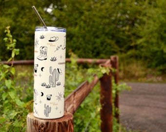 Peut inclure: Un gobelet en acier inoxydable blanc avec un motif noir et blanc représentant des cactus, des bottes de cowboy et le texte "Wild West" et "Country". Le gobelet est posé sur une surface en bois avec une paille en métal qui sort du haut.