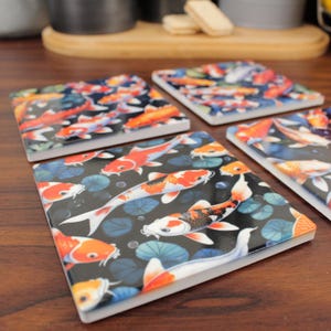 May include: A set of four square coasters with a koi carp design. The coasters display orange, white, and black koi carp swimming amongst lily pads on a dark background. Each coaster is approximately 10 cm square.