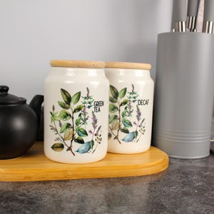 Op de afbeelding: Twee witte keramische potten met houten deksels, één met het opschrift "GREEN TEA" en de andere met "DECAF". Elke pot heeft een botanische illustratie van groene bladeren en paarse bloemen. Een zwarte theepot en een grijze keukengerei houder zijn ook zichtbaar.