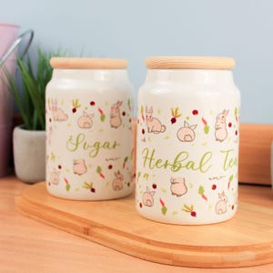 Könnte beinhalten: Zwei weiße Keramikbehälter mit Holzdeckeln, verziert mit Cartoon-Hasen, Karotten und Radieschen. Auf einem Behälter steht "Sugar" und auf dem anderen "Herbal Tea" in grüner Schrift. Die Behälter stehen auf einem Holzschneidebrett.