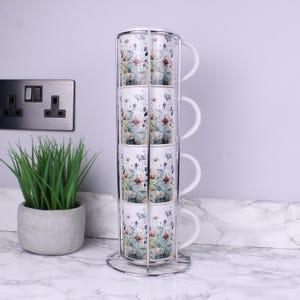 May include: A vertical chrome metal stand holding four stacked ceramic mugs. Each mug features a colourful floral pattern with blue, pink, and green botanical illustrations on a white background. The mugs have matching white handles and rims.