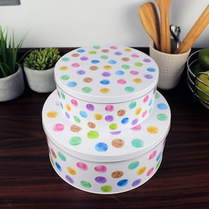 Peut inclure: Deux boîtes rondes blanches pour gâteaux avec un motif de pois aquarelle dans des tons de rose, violet, vert, bleu et marron. Les boîtes sont empilées l'une sur l'autre.
