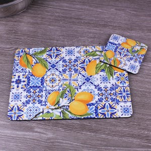 May include: A rectangular placemat and a square coaster featuring a white background with a blue and yellow geometric tile pattern. The design includes illustrations of yellow lemons with green leaves and white blossoms scattered across the surface.