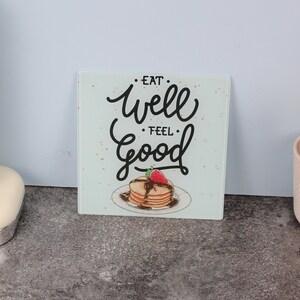 Può includere: Tagliere quadrato in vetro con le parole "Eat Well Feel Good" in un carattere scritto a mano. L'immagine presenta una pila di pancake con sciroppo al cioccolato e una fragola sopra.