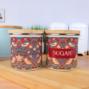 May include: Two ceramic canisters with a floral pattern and a wooden lid. The canister on the right has the word "SUGAR" printed on it.