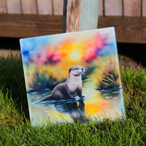 Könnte beinhalten: Ein Glasuntersetzer mit einer farbenfrohen Sonnenuntergangsszene, die ein braunes Otter zeigt, das in einem Gewässer schwimmt. Das Otter schaut nach rechts und der Sonnenuntergang ist im Hintergrund.