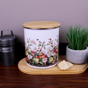 May include: A cylindrical white storage canister with a bamboo lid, featuring a colourful botanical print of apples, green leaves, and white blossoms. The canister is positioned on a bamboo tray next to a dark metal sugar container and a small plant.