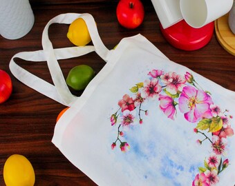 May include: White tote bag with a floral design featuring pink flowers and green leaves on a blue watercolour background. The bag has two white handles.