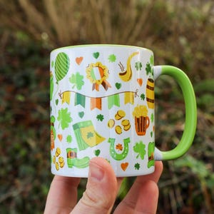 May include: A white ceramic mug with a green rim and handle. The mug is decorated with a pattern of shamrocks, gold coins, and other St. Patrick's Day themed imagery.