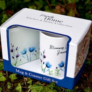 Könnte beinhalten: Ein weißes Geschenkset mit Tasse und Untersetzer mit blauem Blumenmuster. Die Tasse trägt den Text "Blooming Love From". Die Verpackung ist weiß mit blauen Verzierungen und dem Text "Purely Home Kitchen & Home Collection" darauf.