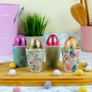 May include: Four colourful ceramic egg cups with different floral and patterned designs. Each cup holds a chocolate egg wrapped in foil, one pink, one gold, and two gold. The egg cups are on a wooden chopping board with small chocolate eggs scattered around.