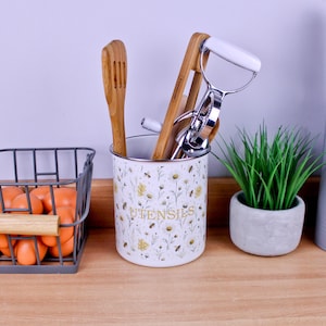 May include: A white cylindrical utensil holder with a floral and bee pattern, featuring the word UTENSILS in gold lettering. It contains a wooden slotted spoon, a wooden tong, and a chrome-plated rotary egg beater with a white handle.