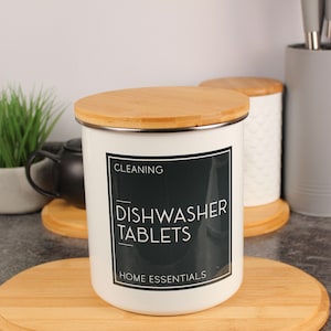 May include: A white cylindrical storage canister with a bamboo lid. The front features a black rectangular label with white text that reads: CLEANING, DISHWASHER TABLETS, HOME ESSENTIALS. The canister has a smooth finish and a metal rim.
