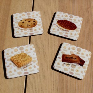 May include: Four square coasters with cookie designs. One coaster features a chocolate chip cookie, another a chocolate digestive, a third a custard cream, and the last a bourbon cream cookie. Each coaster has a white background with cookie illustrations.