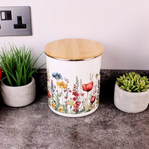 May include: A cylindrical white storage canister featuring a colourful botanical print of various wildflowers including red poppies, yellow blossoms, and blue cornflowers, topped with a circular light-toned wooden lid with a smooth finish.
