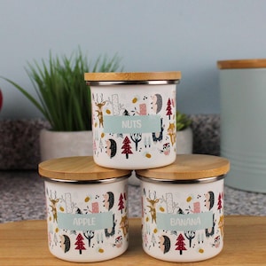 May include: Three white ceramic canisters with bamboo lids and colourful woodland animal illustrations. The canisters are labelled "Nuts", "Apple", and "Banana".