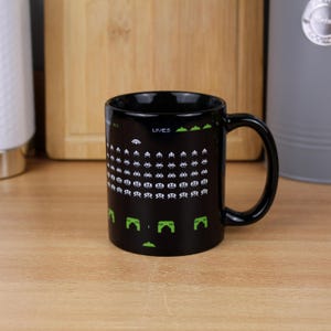May include: A black ceramic mug featuring a retro pixelated video game graphic. The design displays rows of white alien invaders, green defensive bunkers at the bottom, and the text LIVES in a pixelated font against the dark background.