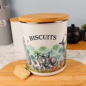 Peut inclure: Pot à biscuits en céramique blanche avec couvercle en bambou. L'illustration en noir et blanc représente des personnages du Magicien d'Oz, avec le mot "BISCUITS" en noir. Deux biscuits sont sur une planche à découper en bois.
