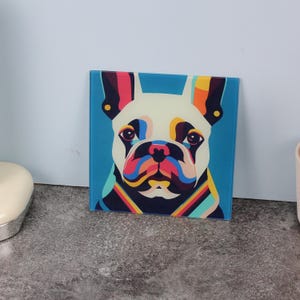 Op de afbeelding: Vierkante glaskunst met een kleurrijk pop-art portret van een Franse Bulldog. Het gezicht van de hond is wit met levendige accenten van blauw, rood, geel en oranje. De achtergrond is lichtblauw. Het kunstwerk wordt op een grijze ondergrond getoond.