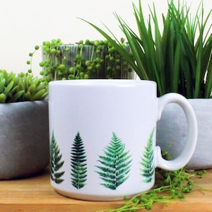 May include: White ceramic mug with a green fern pattern. The mug has a handle and is sitting on a wooden surface with green plants in the background.