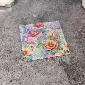 Puede incluir: Posavasos cuadrado de cristal con diseño floral en acuarela. El posavasos muestra una disposición de flores naranjas, rosas, moradas y azules con hojas verdes sobre un fondo claro. Un artículo decorativo para la decoración del hogar.