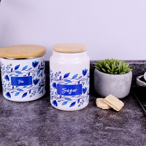 May include: Two white cylindrical canisters with light wood lids, featuring a blue floral vine pattern and blue rectangular labels with the white text "Tea" and "Sugar" printed on them. The canisters are positioned on a dark grey surface.