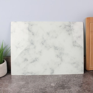May include: A rectangular white marble-effect chopping board with a glossy finish and grey veining. A wooden chopping board is visible on the right. A small potted plant is on the left. The board is suitable for kitchen use.