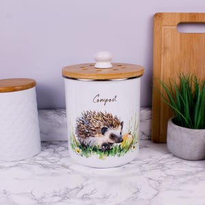 Op de afbeelding: Een witte cilindervormige compostbak met een aquarelillustratie van een egel in groen gras met een gele appel. De container heeft een lichtbruin houten deksel met een witte knop en ventilatiegaten, en het woord Compost staat erop gedrukt.