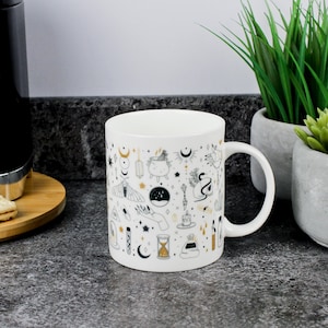 May include: White ceramic mug featuring a wraparound design of celestial and mystical symbols in black and gold. The pattern includes moons, stars, hands, potions, and other magical elements. The mug has a curved handle and is set against a neutral background.