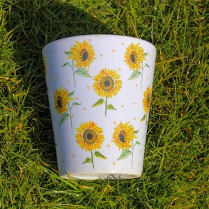 May include: White ceramic planter with a sunflower pattern. The planter has a wide mouth and a rounded bottom. It is decorated with yellow sunflowers and small white dots.