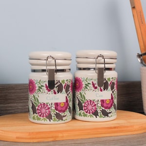 Op de afbeelding: Twee witte keramische potten met bloemen- en dassenontwerpen. Eén leest "Coffee" en de andere "Tea". Elk heeft een crèmekleurig deksel en een metalen sluiting. De potten staan op een houten snijplank.