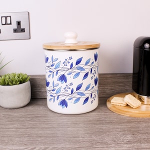 May include: White ceramic canister with a bamboo lid and a white knob. The canister is decorated with a blue floral pattern. The canister is on a wooden surface.