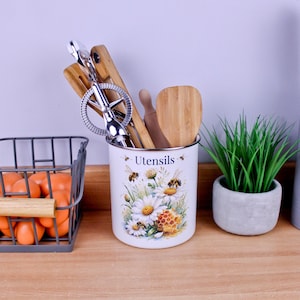 May include: A white cylindrical metal utensil holder featuring a floral print of daisies, honeycombs, and bees with the text "Utensils" printed on the front. The container is filled with various wooden kitchen tools and a silver metal whisk.