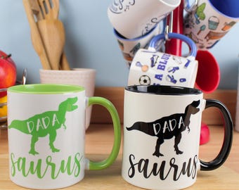 Può includere: Due tazze in ceramica bianca con grafica di dinosauri e la scritta "Dada Saurus". Una tazza ha l'interno e il manico verdi, l'altra ha l'interno e il manico neri. Altre tazze sono visibili sullo sfondo.