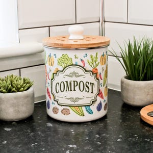 Könnte beinhalten: Weißer Komposteimer mit Bambusdeckel und weißem Knauf. Der Behälter ist mit farbenfrohen Illustrationen von Gemüse und dem Wort "COMPOST" in einem dekorativen Rahmen verziert. Auf einer schwarzen Arbeitsplatte.