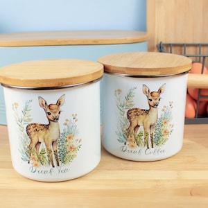 May include: Two white metal canisters with wooden lids. Each canister features a watercolour illustration of a fawn surrounded by flowers. One reads "Decaf Tea" and the other "Decaf Coffee". The canisters are designed for kitchen storage.