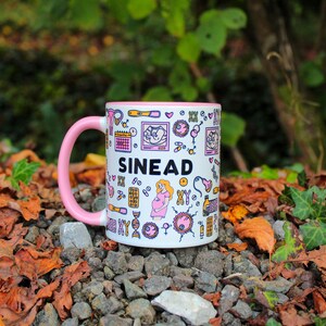 May include: A white ceramic mug with a pink rim and handle. The mug has a black and white design with the name "SIN&Eacute;AD" printed on it. The design includes various illustrations of medical symbols, including a stethoscope, a DNA strand, and a heart.