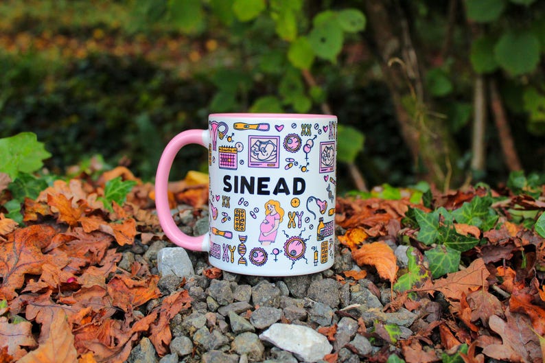 May include: A white ceramic mug with a pink rim and handle. The mug has a black and white design with the name "SIN&Eacute;AD" printed on it. The design includes various illustrations of medical symbols, including a stethoscope, a DNA strand, and a heart.