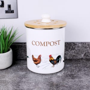 May include: A white cylindrical compost bin with a metal body and a light wood lid featuring a white knob. The bin displays the word COMPOST in brown text above a colourful illustration of a hen and a rooster in a rural landscape setting.