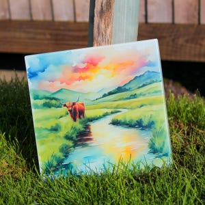 Puede incluir: Una pintura de acuarela sobre una tabla de cortar de vidrio representa una vaca Highland roja de pie en un campo verde con un arroyo que la atraviesa. El cielo es una puesta de sol naranja y rosa vibrante.