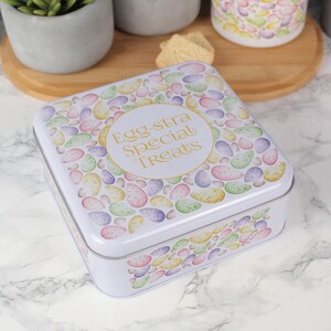 May include: A white, square tin with a lid decorated with a pattern of pastel-colored, egg-shaped designs. The centre of the lid features a gold circle with the words "Egg-stra Special Treats". The tin is likely for storing treats.