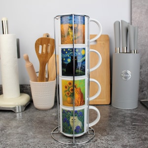 May include: A set of four stacked ceramic mugs with cat-themed artwork, held in a chrome metal rack. Each mug features a different cat-themed painting, including a cat in a field, a black cat under a starry night, sunflowers, and a white cat in a garden.